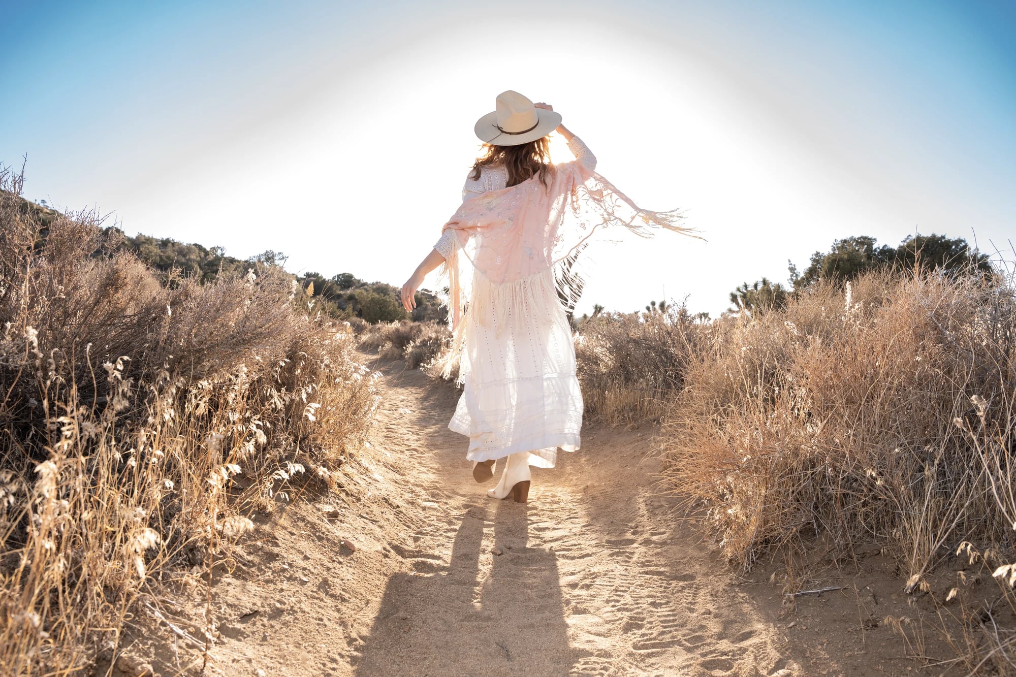 Shannon walking a sunlit high-desert path in a flowing white dress